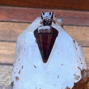 Puede incluir: Un colgante de forma triangular en color marrón oscuro con un acabado liso y pulido, y una tapa metálica con intrincados patrones decorativos en espiral. El colgante cuelga de una cadena color cobre y descansa sobre un cristal claro y dentado.