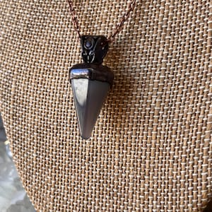 Puede incluir: Un collar con colgante que presenta una punta de piedra gris, lisa y triangular, suspendida de una tapa metálica oscura con detalles de filigrana. El colgante cuelga de una cadena metálica fina de color cobre, creando un contraste entre el metal oscuro y la piedra gris clara.