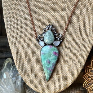 May include: A handcrafted necklace featuring a large, teardrop-shaped pendant with a green and red stone. The pendant is accented with smaller stones and a decorative metalwork setting. The necklace hangs on a delicate copper chain.