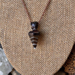 Puede incluir: Un collar con colgante que presenta una piedra cónica con bandas marrones y blancas suspendida de una cadena de metal color cobre. La piedra tiene un acabado liso y pulido con rayas horizontales, y la tapa de metal tiene un acabado oscuro y texturizado.