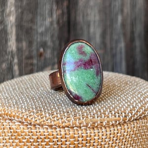 May include: A copper-colored ring featuring a large, oval-shaped stone. The stone has a vibrant green base color with striking red inclusions. The ring is displayed on a tan, woven surface, showcasing the jewelry's unique design.