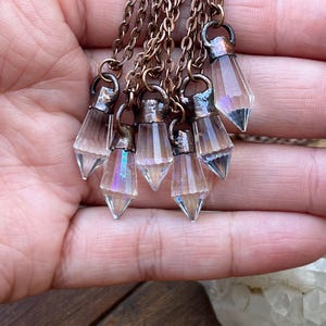 Puede incluir: Un conjunto de colgantes de cristal con facetas iridiscentes, suspendidos de cadenas de color cobre. Cada colgante es un cristal claro y facetado con una base puntiaguda. Las cadenas están unidas a una pequeña tapa metálica.