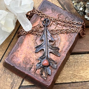 May include: A copper necklace with an oxidized silver oak leaf pendant and a red carnelian stone. The necklace is displayed on a copper-colored surface.