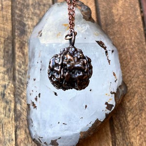 May include: A bronze-colored pendant necklace featuring a textured, spherical charm. The charm hangs from a delicate copper-colored chain. The necklace is displayed against a large, clear crystal with a wooden background.