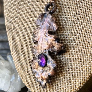 May include: A handcrafted copper pendant necklace featuring an oak leaf design. The leaf pendant has a dark patina and a faceted purple gemstone. The necklace is displayed on a burlap-textured surface.