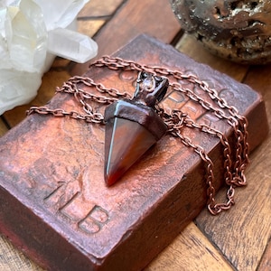 Puede incluir: Un colgante triangular puntiagudo con una piedra de color marrón rojizo oscuro engastada en una tapa de metal de tono cobre oxidado oscuro. El colgante cuelga de una cadena de metal de tono cobre a juego, sobre un bloque de cobre marcado 1LB.