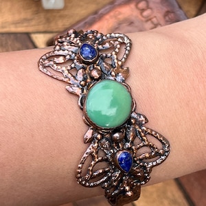 May include: A copper-toned cuff bracelet with an openwork design. The bracelet features a large, round, light green stone in the center, flanked by two smaller, blue stones. The intricate metalwork has a rustic, antique appearance.