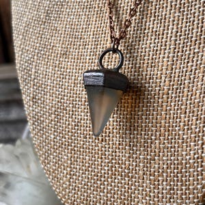 Puede incluir: Un collar con colgante que presenta una piedra triangular translúcida con una tapa de metal oscuro. El colgante cuelga de una delicada cadena de color cobre. El collar se exhibe sobre una tela texturizada de color beige.