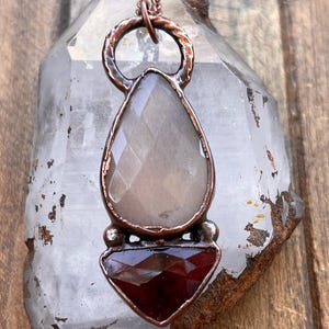 May include: A pendant necklace featuring a faceted, pear-shaped gray stone positioned above a faceted, triangular deep red stone. Both stones are encased in a textured, dark copper-toned metal setting with a circular loop at the top.