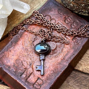 May include: A key pendant necklace with a dark oval stone and a copper chain. The key is a dark silver color. The necklace rests on a textured, reddish-brown surface. A clear crystal and a metallic object are in the background.