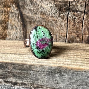 May include: Oval-shaped ring featuring a green and red gemstone set in a copper-colored metal band. The gemstone has a speckled pattern with a mix of green and red hues. The ring is displayed on a wooden surface.