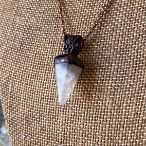 Puede incluir: Un collar con un colgante de piedra blanca translúcida de forma triangular, rematado con un engaste de metal oscuro y texturizado. El colgante cuelga de una fina cadena de metal color cobre, mostrando una forma geométrica puntiaguda.