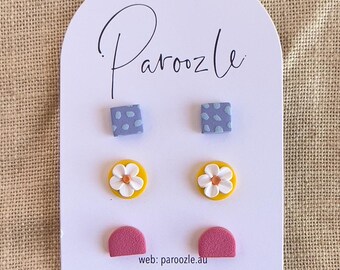 Puede incluir: Un conjunto de seis pendientes de arcilla polimérica de colores. Los pendientes se muestran en una tarjeta blanca con el texto "Paroozle" y la información de contacto. Los pendientes incluyen dos pendientes cuadrados con un patrón azul y blanco, dos pendientes redondos con un fondo amarillo y una flor blanca con un centro naranja, y dos pendientes de medio círculo de color rosa.