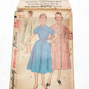 May include: Vintage Simplicity printed sewing pattern envelope. The envelope features illustrations of women's dresses and robes. The main dress is a light blue button-down dress. The envelope is aged with the text "SIMPLE TO MAKE".