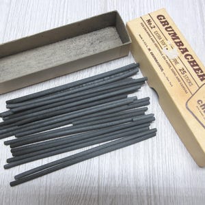 May include: A box of Grumbacher No. 2 Extra Soft artists' charcoal containing 25 sticks for drawing and sketching on paper, signs, and canvas. The set includes a rectangular cardboard box with a separate lid and a collection of thin, dark gray charcoal sticks.