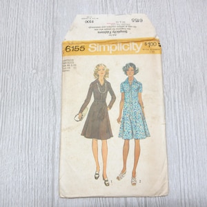 May include: A vintage Simplicity sewing pattern envelope, number 6155, featuring illustrations of two dress styles. One is a long-sleeved solid dress, and the other is a short-sleeved floral dress. The envelope includes text indicating sizes 44 to 50.