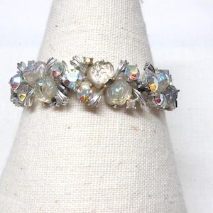 May include: A silver-toned bracelet with clear and iridescent rhinestones. The bracelet features a floral design with multiple clear, round, and teardrop-shaped stones. The rhinestones have an iridescent sheen, reflecting multiple colors.