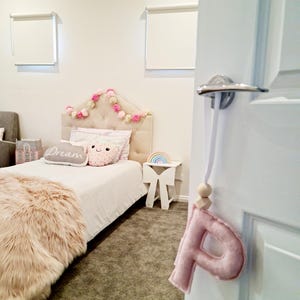 Puede incluir: Una letra P de fieltro rosa colgada de una puerta blanca con una cuenta de madera y una cuerda blanca. La letra está colgada en la habitación de un niño con una cama, una alfombra y una mesita de noche.
