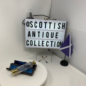Puede incluir: Una caja de luz rectangular con el texto negro @SCOTTISH ANTIQUE COLLECTION, una pequeña bandera escocesa sobre un soporte negro y un frasco de perfume de vidrio transparente con tapas de metal dorado en un estuche marrón abierto.