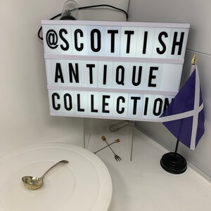 Puede incluir: Una caja de luz rectangular blanca muestra el texto negro @SCOTTISH ANTIQUE COLLECTION. Debajo, una pequeña cuchara colador de plata descansa sobre una base circular blanca. Se ven dos finos palillos de cóctel de metal con puntas de colores.