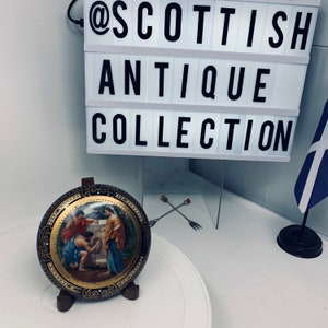 May include: A circular decorative plate featuring a classical scene of three figures in robes, with a gold-toned centre and a black border decorated with gold filigree. Above the plate, a light box displays the text @SCOTTISH ANTIQUE COLLECTION.