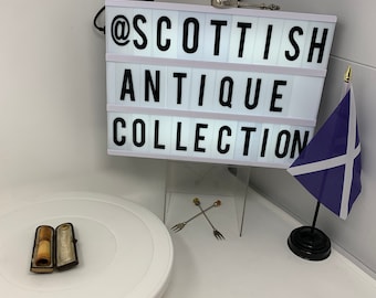 May include: A rectangular light box displaying the text @SCOTTISH ANTIQUE COLLECTION in black letters. Below it sits a small open dark brown case containing a cylindrical object, two thin metal forks with amber-colored tips, and a Scottish flag.