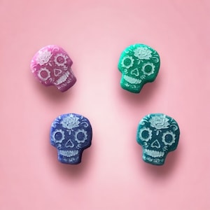 May include: Four sugar skull shaped candy decorations. Two are blue, one is pink, and one is green.