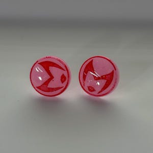 May include: A pair of pink stud earrings with a red glitter design. The design is a stylised letter "M" with a star inside.