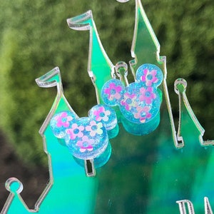 May include: Two sparkly blue and pink Mickey Mouse ears with a floral design, sitting on a green and blue plastic castle.