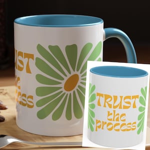 May include: White ceramic mug with a blue interior and handle. The mug features a large green and yellow daisy design with the words "TRUST the process" in yellow lettering. The mug is sitting on a wooden surface.