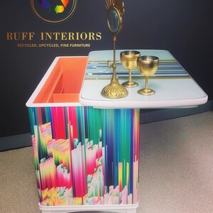 May include: A white bar cart with a colorful abstract design and a white top. The cart has a gold mirror and two gold goblets on the top. The cart is on wheels and has a small drawer. The cart is labeled "RUFF INTERIORS, RECYCLED, UPCYCLED, FINE FURNITURE".