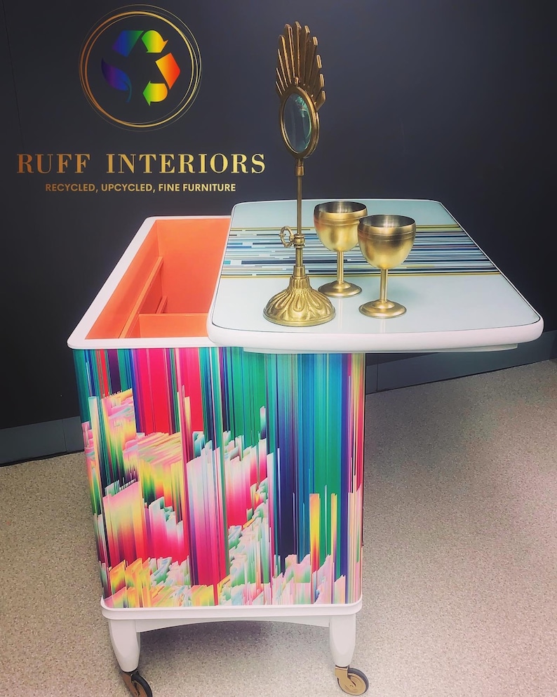 May include: A white bar cart with a colorful abstract design and a white top. The cart has a gold mirror and two gold goblets on the top. The cart is on wheels and has a small drawer. The cart is labeled "RUFF INTERIORS, RECYCLED, UPCYCLED, FINE FURNITURE".
