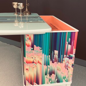 May include: A white side table with a lift-top and a colorful abstract design on the front. The table has a light blue top and a pink interior. The table is on wheels.