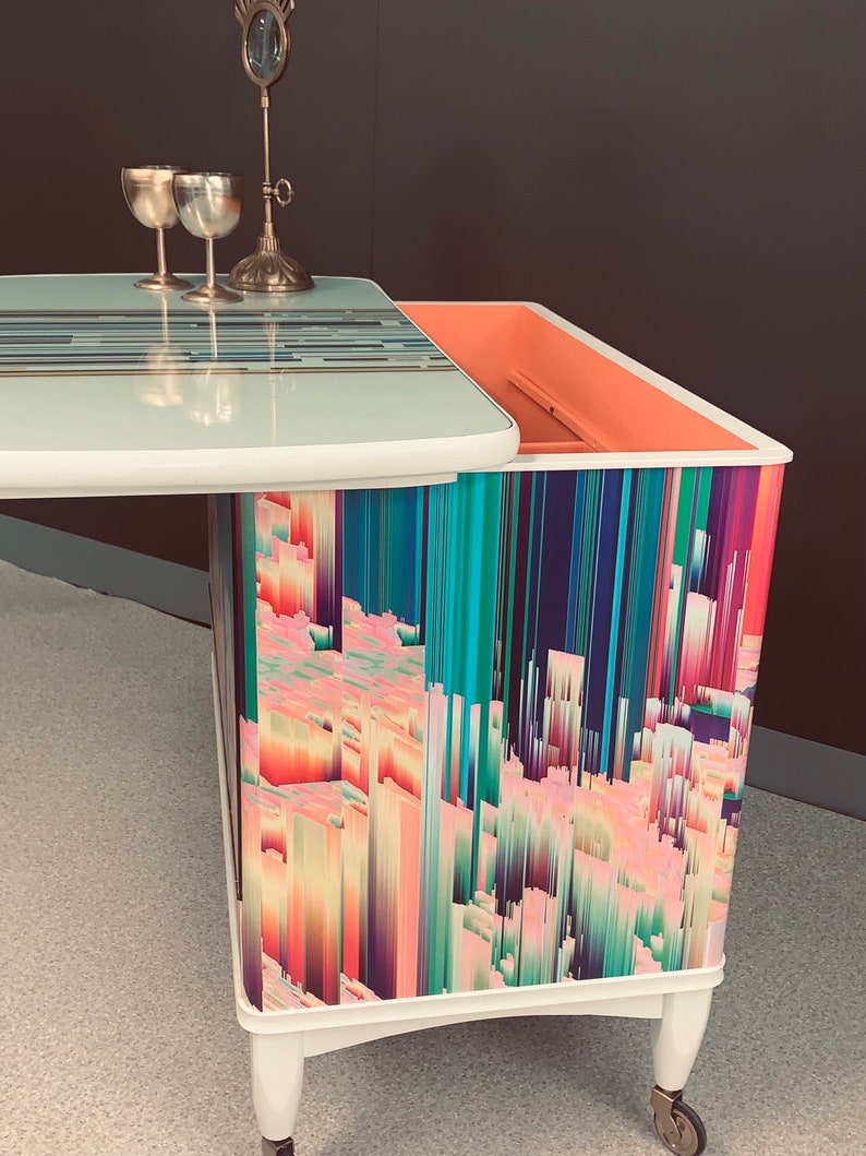 May include: A white side table with a lift-top and a colorful abstract design on the front. The table has a light blue top and a pink interior. The table is on wheels.
