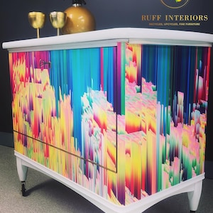 May include: A white cabinet with a colorful abstract design. The cabinet has a single door and two drawers. It is on wheels and has a gold trim. The cabinet is labeled "RUFF INTERIORS RECYCLED, UPCYCLED, FINE FURNITURE".