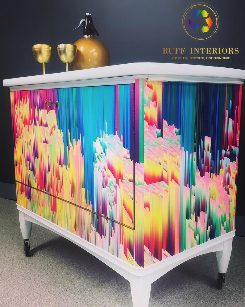 May include: A white cabinet with a colorful abstract design. The cabinet has a single door and two drawers. It is on wheels and has a gold trim. The cabinet is labeled "RUFF INTERIORS RECYCLED, UPCYCLED, FINE FURNITURE".