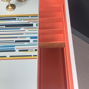 May include: A white bar cabinet with a geometric pattern on the top and a coral-colored interior with four shelves. The cabinet is open and the shelves are empty.