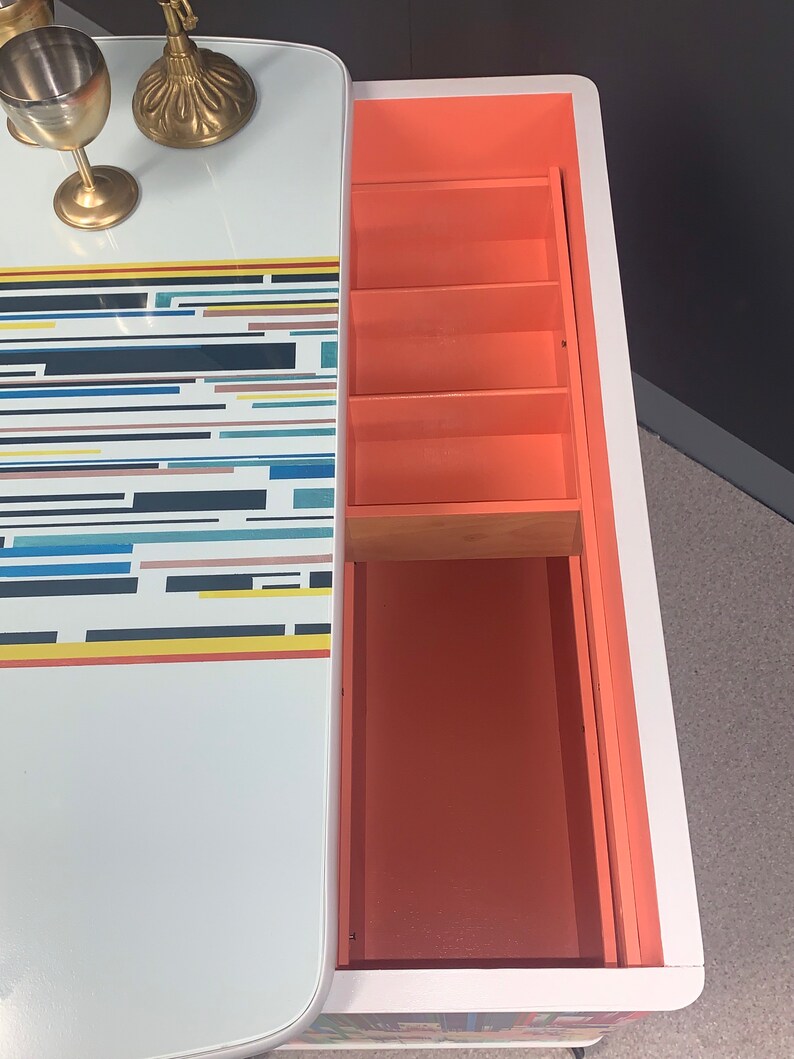 May include: A white bar cabinet with a geometric pattern on the top and a coral-colored interior with four shelves. The cabinet is open and the shelves are empty.