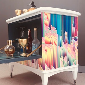 May include: White bar cart with a colorful abstract design on the front. The cart has a drop-down door and is on wheels. It is filled with glassware and bottles.