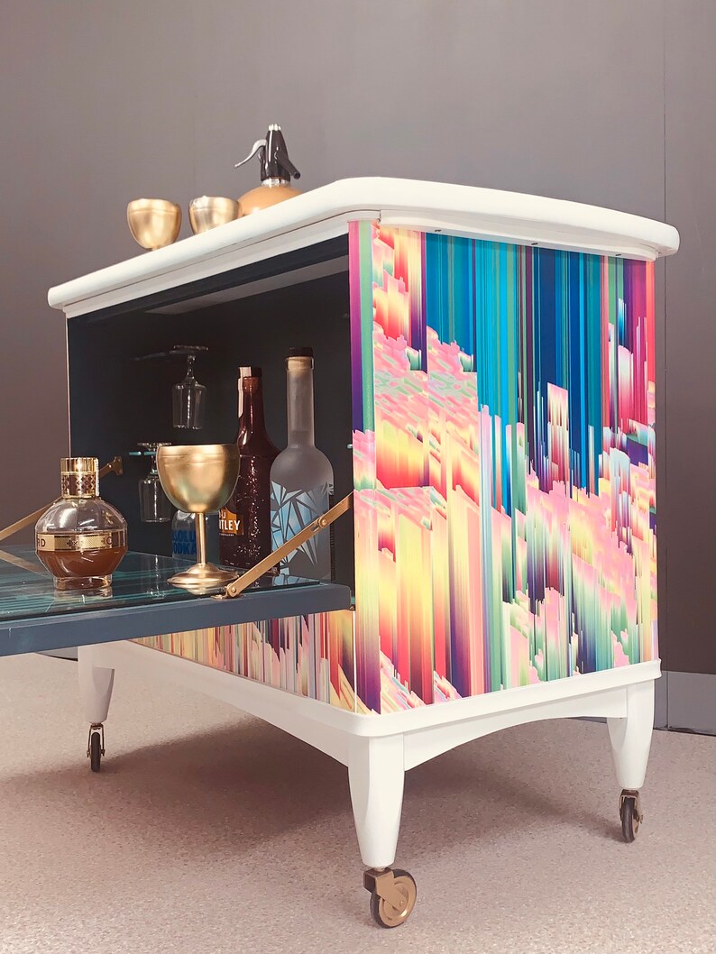 May include: White bar cart with a colorful abstract design on the front. The cart has a drop-down door and is on wheels. It is filled with glassware and bottles.