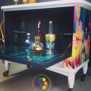 May include: A white bar cart with a black interior and a glass top. The cart has a rainbow patterned side panel and gold accents. The cart is on wheels and has a bottle of Absolut Vodka and a bottle of liquor inside.