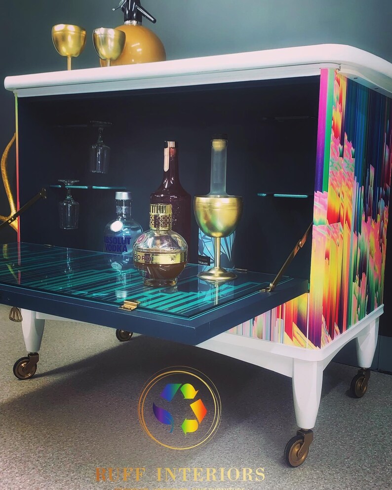 May include: A white bar cart with a black interior and a glass top. The cart has a rainbow patterned side panel and gold accents. The cart is on wheels and has a bottle of Absolut Vodka and a bottle of liquor inside.
