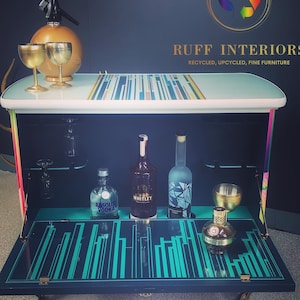 May include: A turquoise and black bar cart with a mirrored top and a striped design. The cart has a fold-down front and is on wheels. The cart is stocked with liquor bottles and glasses. The cart is labeled "RUFF INTERIORS RECYCLED, UPCYCLED, FINE FURNITURE".