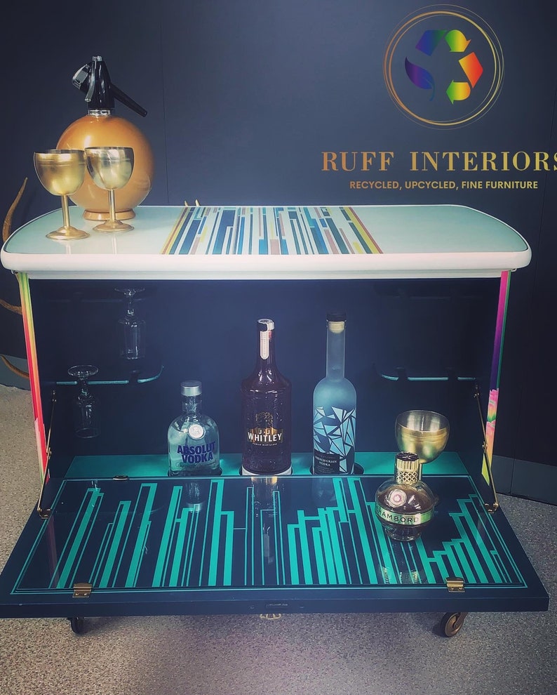 May include: A turquoise and black bar cart with a mirrored top and a striped design. The cart has a fold-down front and is on wheels. The cart is stocked with liquor bottles and glasses. The cart is labeled "RUFF INTERIORS RECYCLED, UPCYCLED, FINE FURNITURE".