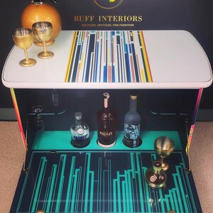 May include: A white bar cart with a colorful geometric design on the top. The cart has a fold-down front with a turquoise and black geometric design. The cart is open and has three bottles of liquor and a glass on the shelf. The cart is labeled "RUFF INTERIORS, RECYCLED, UPCYCLED, FINE FURNITURE".