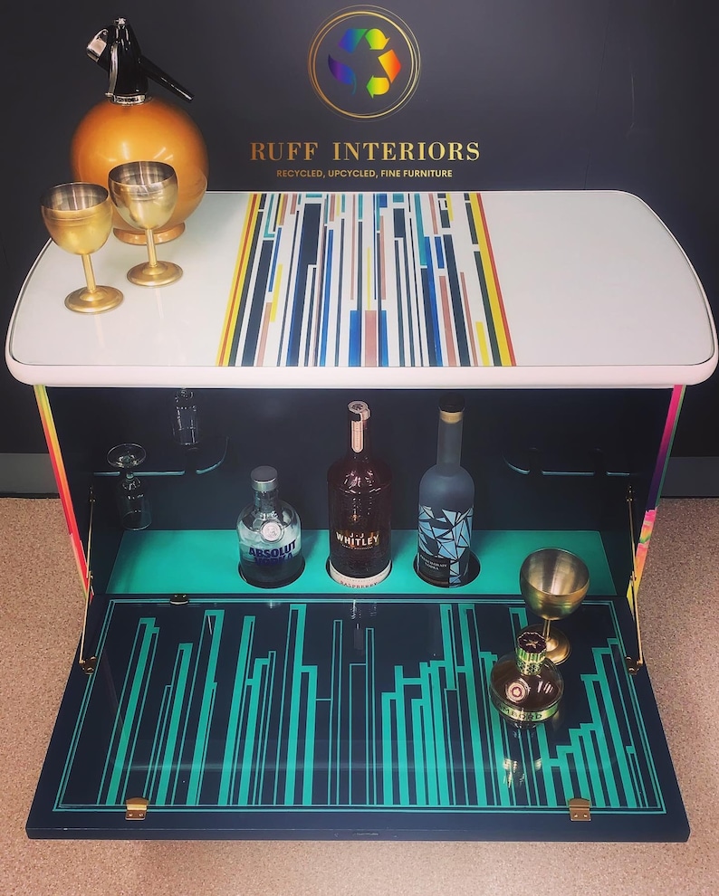 May include: A white bar cart with a colorful geometric design on the top. The cart has a fold-down front with a turquoise and black geometric design. The cart is open and has three bottles of liquor and a glass on the shelf. The cart is labeled "RUFF INTERIORS, RECYCLED, UPCYCLED, FINE FURNITURE".