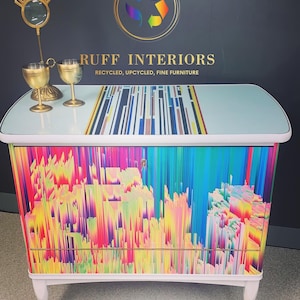 May include: A white cabinet with a colorful abstract design on the front. The cabinet has two drawers and is on wheels. The top of the cabinet is light blue with a black and white striped design. The text "RUFF INTERIORS RECYCLED, UPCYCLED, FINE FURNITURE" is on the top of the cabinet.