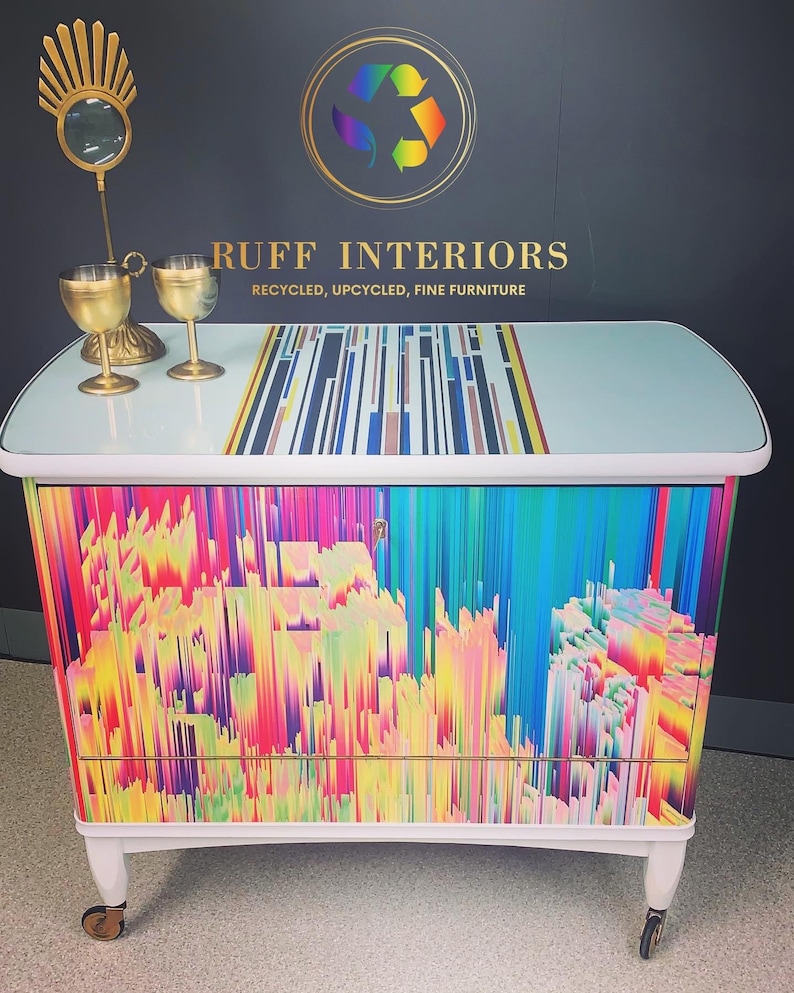 May include: A white cabinet with a colorful abstract design on the front. The cabinet has two drawers and is on wheels. The top of the cabinet is light blue with a black and white striped design. The text "RUFF INTERIORS RECYCLED, UPCYCLED, FINE FURNITURE" is on the top of the cabinet.