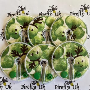 May include: Six green and white fabric breast pads with a reindeer design. The pads have a hole in the centre and a green button on the side.