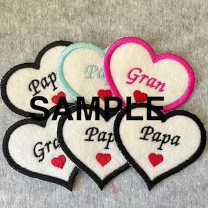 May include: Five felt heart-shaped patches with black stitching around the edges. The patches are white with black embroidered text. The text on the patches reads "Pap", "Pa", "Gran", "Pap" and "Papa". Each patch has a small red heart embroidered in the bottom right corner.